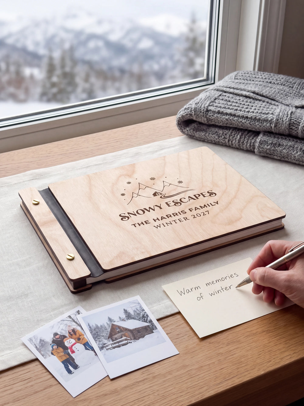 A Natural wooden album showing snowy mountains, a cabin and snowflakes rests beside a separate gift card. A hand writes 