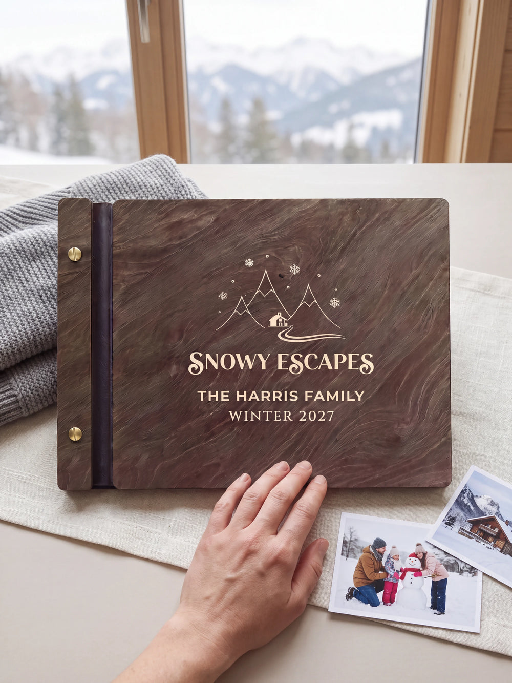 A hand rests beside the closed dark brown Jacobean album on a table. Cream engraving and personalized wording are visible, with travel photos nearby.