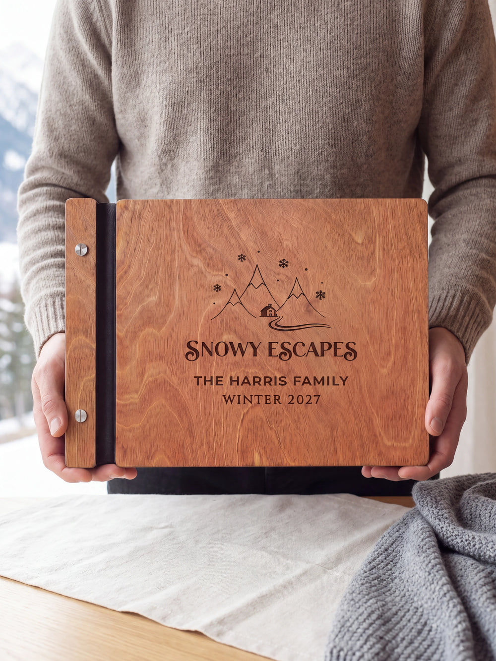 Hands hold the Hazelnut wooden album near a window. The full cover displays snowy mountains, a cabin and snowflakes in dark engraving, with personalized lettering and a black binding strip.