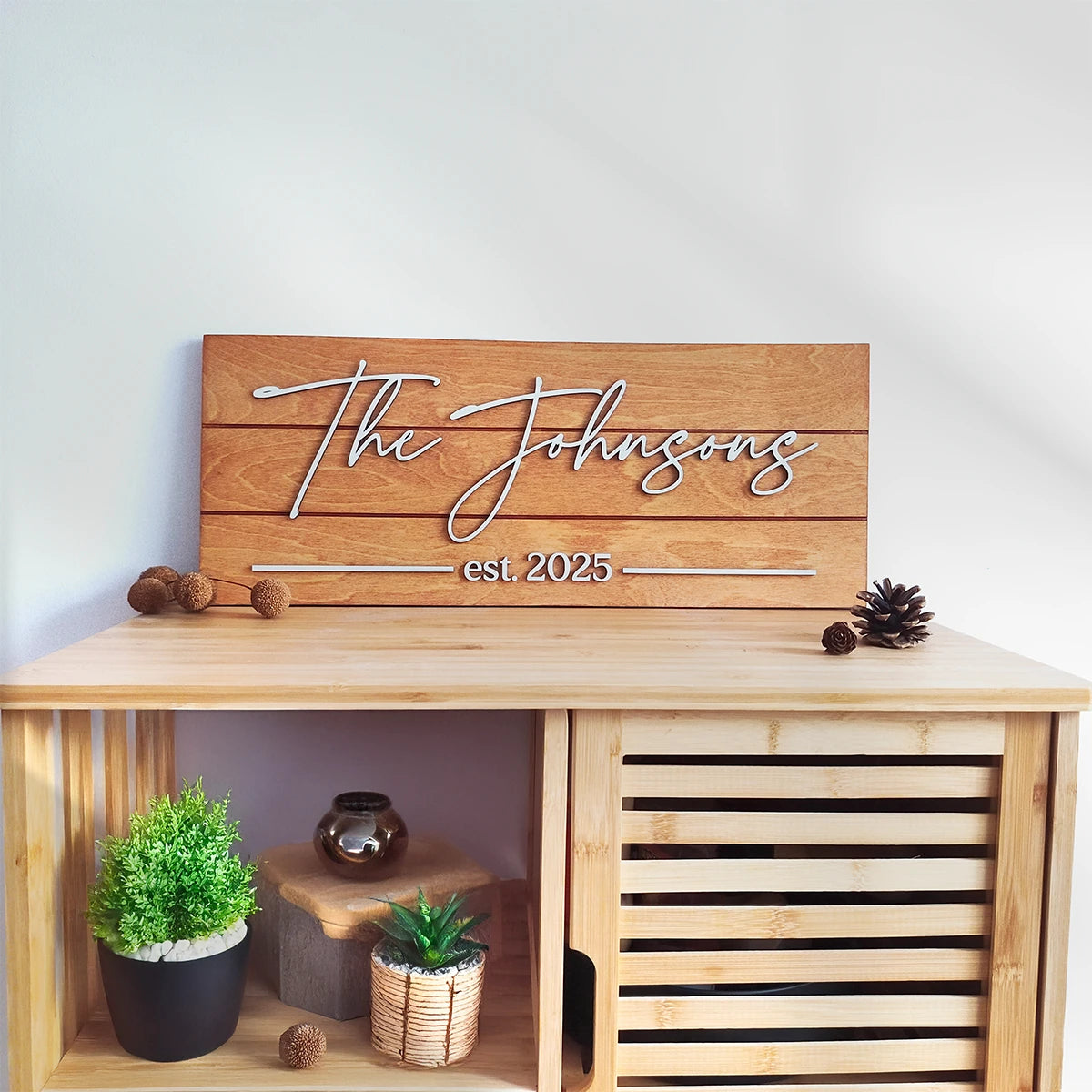 Wooden sign with 'The Johnsons est 2025' on a wooden shelf with decorative items.