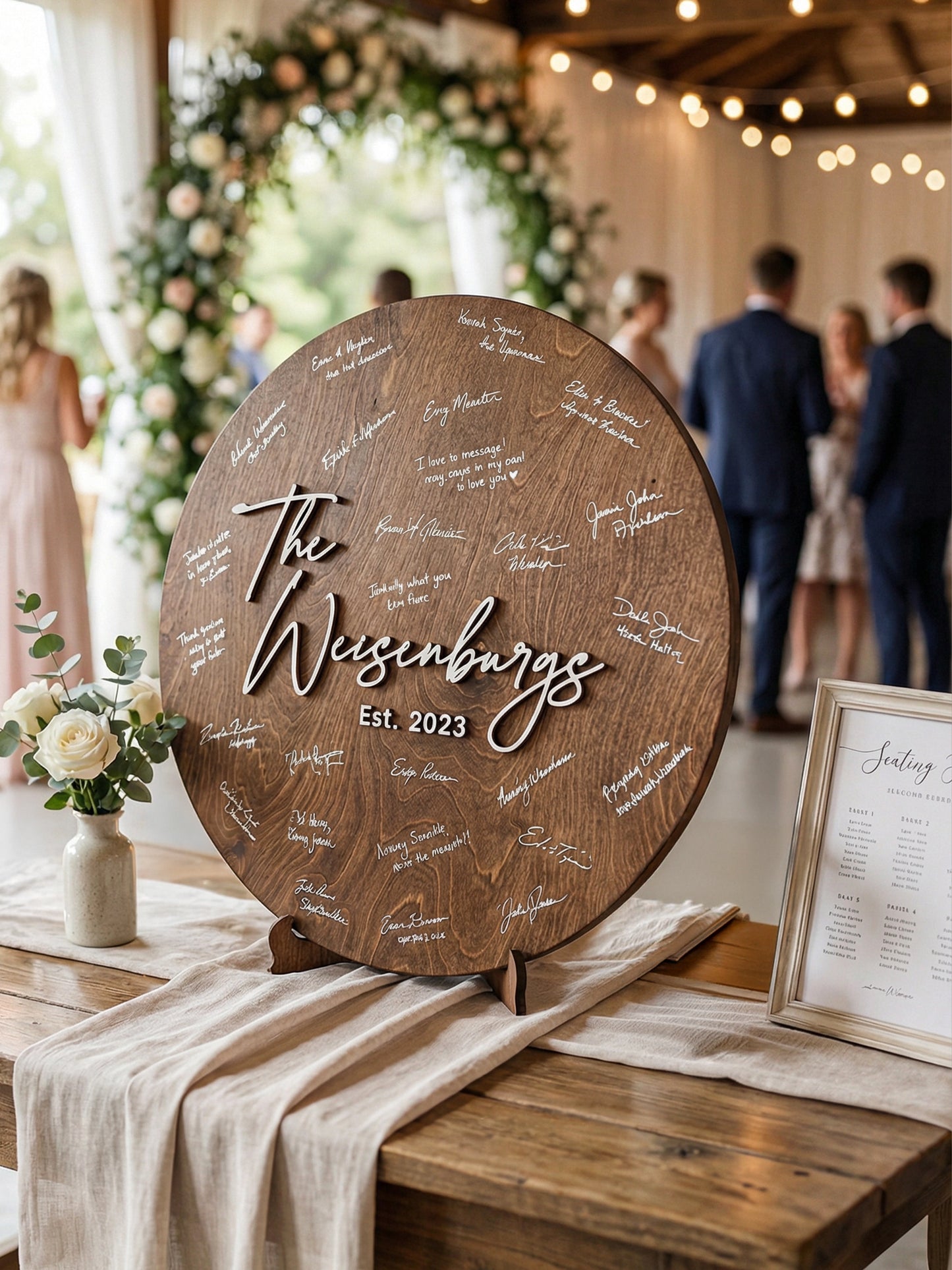 Wooden round sign with engraved text on a table with floral decorations and people in the background